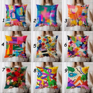 May include: Nine decorative throw pillows with colorful abstract designs. Each pillow features a unique pattern, including geometric shapes, floral motifs, and vibrant color palettes. The pillows are square-shaped and held by a person.