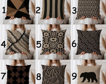 May include: Nine decorative throw pillows with various geometric and nature-inspired designs. Pillows feature patterns like triangles, stripes, waves, flowers, and a bear silhouette. Colors include black, brown, and beige, offering a range of styles for home decor.