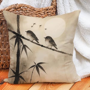Op de afbeelding: Vierkant kussen met een aquarel schilderij van twee vogels op een bamboetak. Het ontwerp toont een volle maan en vliegende vogels tegen een zachte, neutrale achtergrond. Het kleurenpalet omvat beige, zwart en grijstinten.