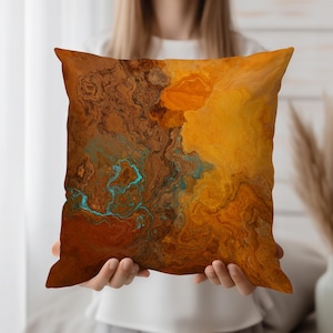 May include: Square cushion with an abstract design in shades of orange, brown, and teal. The cushion features a marbled or fluid art pattern, creating a textured and artistic appearance. The cushion is held in front of a person.