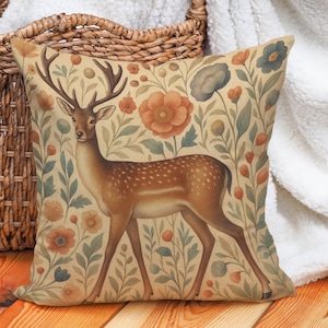 Könnte beinhalten: Quadratisches Zierkissen mit einem Hirschmotiv, umgeben von Blumen und Blättern in Orange-, Blau- und Grüntönen. Der Hintergrund ist hellbeige. Das Kissen befindet sich in der Nähe eines Weidenkorbs und einer weißen Decke.