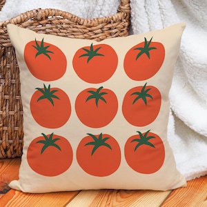 May include: Square throw pillow with a cream background featuring a pattern of nine large, red tomatoes with green stems. The pillow is placed in front of a woven basket and a white blanket.
