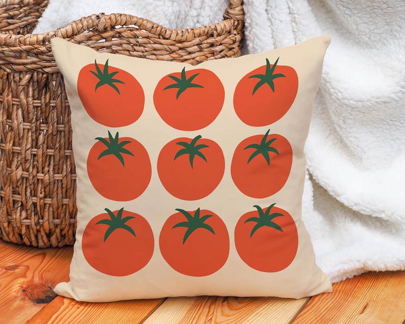 May include: Square throw pillow with a cream background featuring a pattern of nine large, red tomatoes with green stems. The pillow is placed in front of a woven basket and a white blanket.