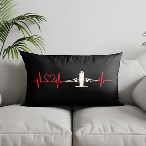May include: Black rectangular pillow featuring a red heartbeat line with a heart shape and a white airplane graphic. The pillow is on a white couch with a green plant in the background.