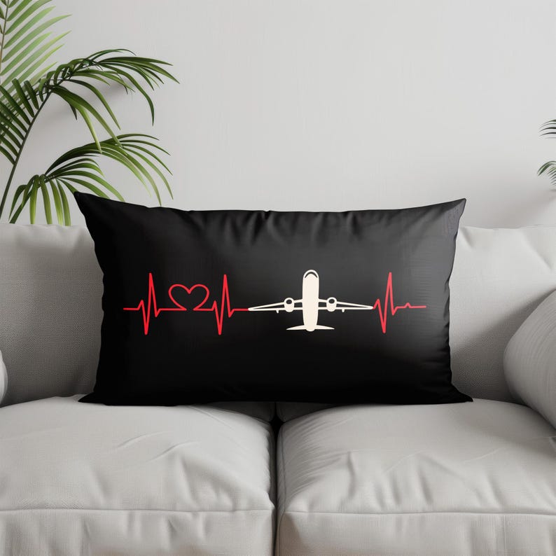 May include: Black rectangular pillow featuring a red heartbeat line with a heart shape and a white airplane graphic. The pillow is on a white couch with a green plant in the background.