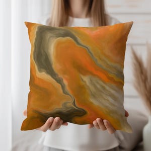May include: Square decorative pillow with an abstract design in shades of orange, brown, and cream. The pillow features a swirling, marbled pattern, creating a fluid and artistic appearance. The pillow is held in front of a person.