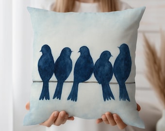 May include: A square decorative pillow featuring a watercolor design of five navy blue birds perched on a thin wire. The birds are silhouetted against a light blue and white background. The pillow is held by a person.