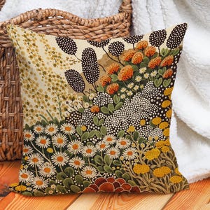 Op de afbeelding: Vierkant sierkussen met een bloemenontwerp. Het kussen heeft een kleurrijk patroon van madeliefjes, oranje bloemen en andere botanische elementen. De achtergrond is een mix van crème, bruin en zwart. Het kussen ligt op een houten oppervlak.