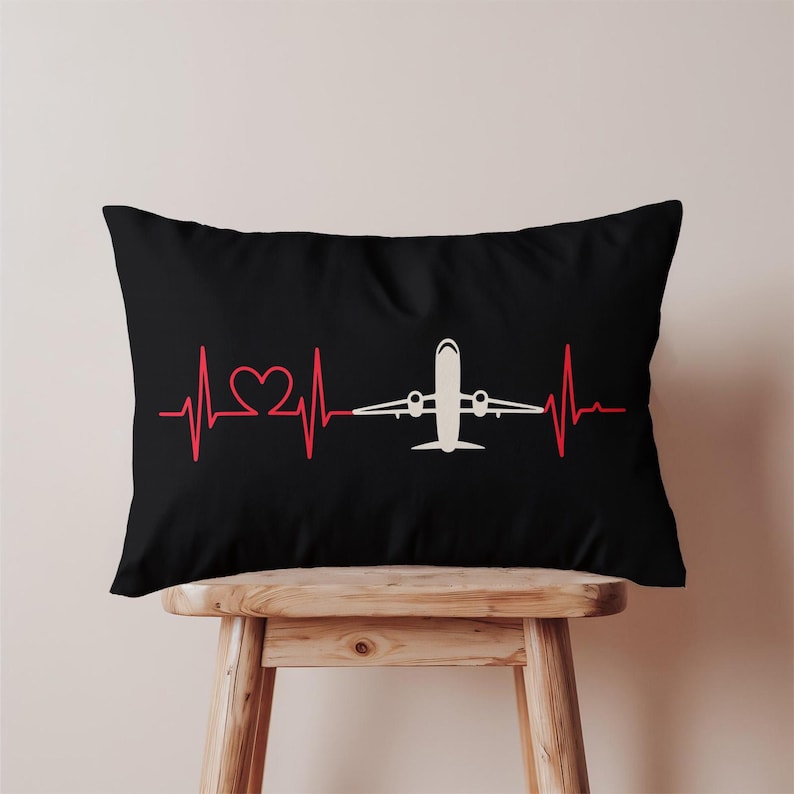 May include: Black rectangular pillow featuring a red heartbeat line with a heart shape and a white airplane graphic. The pillow is resting on a wooden stool. The design is simple and graphic.