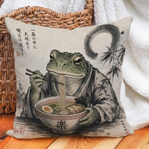 Op de afbeelding: Een vierkant sierkussen met een bedrukte illustratie van een groene kikker in een kimono, die met stokjes ramen eet uit een kom. Het ontwerp bevat zwarte inktstreken, bamboestengels en Japanse teksttekens.