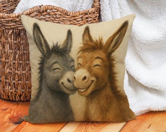 May include: A decorative pillow featuring a whimsical illustration of two smiling donkeys. One donkey is gray, the other is brown, and they are nuzzling each other. The pillow has a neutral background and is placed near a wicker basket.