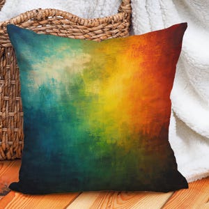 Peut inclure: Coussin carré avec un motif abstrait. Le coussin présente un dégradé de couleurs, passant du bleu foncé au vert, jaune, orange et rouge. Le coussin est posé sur une surface en bois à côté d'un panier en osier et d'une couverture blanche.