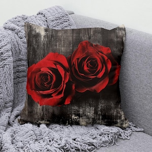 Op de afbeelding: Decoratief sierkussen met twee levendige rode rozen tegen een verweerde, donkere houtnerf achtergrond. Het vierkante kussen ligt op een grijze bank, met een grijze gebreide deken over de arm.