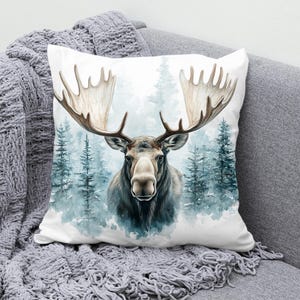 May include: A white cushion featuring a watercolour illustration of a moose in a snowy woodland setting. The moose has large antlers and is looking directly at the viewer.