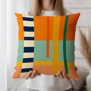 Peut inclure: Coussin carré avec un motif géométrique abstrait. Le coussin présente une palette de couleurs vibrantes d'orange, de sarcelle, de jaune et de vert. Un motif à rayures verticales bleu marine et crème est sur le côté gauche. Le coussin est tenu par une personne.