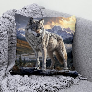 May include: A cushion with a detailed illustration of a grey wolf standing on a log. The background shows a mountain landscape with a sunset, creating a nature-themed design. The cushion is placed on a grey sofa with a knitted throw.