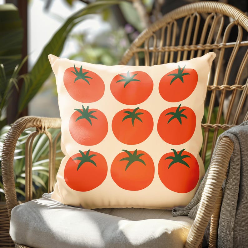 May include: A square decorative pillow with a cream-colored background. The pillow features a pattern of nine large, red tomatoes with green stems. The pillow is placed on a wicker chair with a gray cushion.