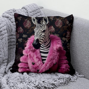 Könnte beinhalten: Dekoratives Zierkissen mit einem Zebra, das einen leuchtend pinkfarbenen Kunstfellmantel trägt. Der Kopf und der gestreifte Körper des Zebras sind vor einem dunklen floralen Hintergrund platziert. Das Kissen liegt auf einem grauen Sofa mit einer passenden Decke.