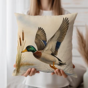 May include: A decorative square pillow featuring a watercolor painting of a mallard duck in flight. The duck has a green head, brown wings, and a yellow beak. The background shows water, reeds, and a light blue sky. The pillow is a neutral beige color.