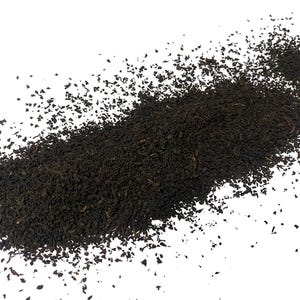 May include: Loose black tea leaves scattered on a white background. The tea leaves are dark brown and have a slightly rough texture.