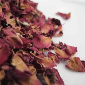 May include: Dried rose petals in shades of pink and red, scattered on a white surface. The petals are delicate and have a slightly wrinkled texture.