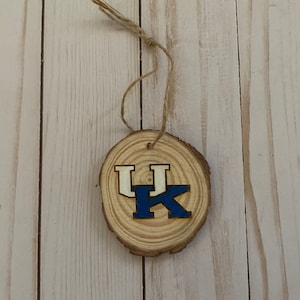 Op de afbeelding: Een ronde houten ornament met een natuurlijke houtnerf. Het heeft de letters "UK" in blauw en wit, met een touwlus om op te hangen. Het ornament heeft een rustieke uitstraling.