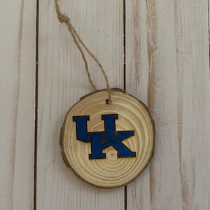 May include: A wooden ornament with a natural wood grain finish. The ornament features a blue and gold logo, suspended by a twine string. The logo is a stylized "UK" design, likely representing a university or team.