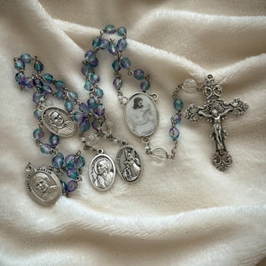 May include: A silver rosary with blue and clear glass beads. The rosary features a silver crucifix and several silver medallions with images of saints. The medallions have the names of the saints inscribed on them, including "Saint Zelie Martin".