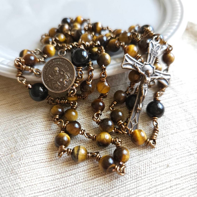 May include: A rosary featuring tiger eye beads, black beads, and a silver crucifix. The rosary also includes a silver medallion with a religious image. The beads are strung on a copper-toned wire.