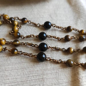 May include: Close-up of three strands of beaded necklaces. The beads alternate between round, polished black stones and tiger's eye stones. The beads are strung on a copper-coloured wire.