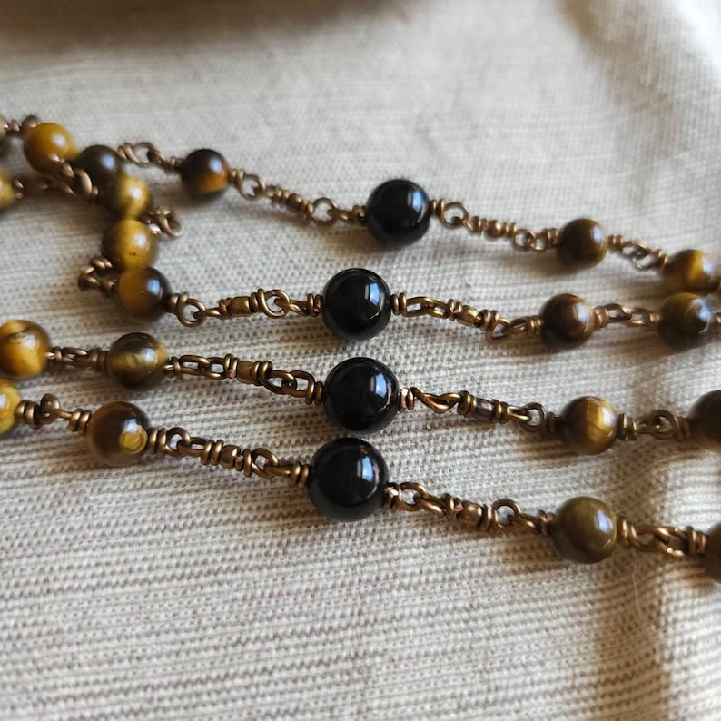 May include: Close-up of three strands of beaded necklaces. The beads alternate between round, polished black stones and tiger's eye stones. The beads are strung on a copper-coloured wire.