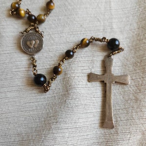 May include: A rosary featuring tiger's eye beads and a silver cross. The beads are a mix of brown and black, strung on a gold-coloured wire. A silver medallion with two hearts is also present.