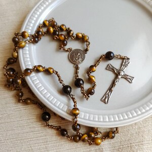 May include: A rosary featuring tiger eye beads and dark brown beads, linked by copper-coloured wire. It includes a silver-coloured crucifix and a medallion with a religious figure. The beads are arranged on a white plate.