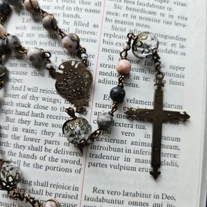 Pu&ograve; includere: Un rosario con una croce d'argento e un ciondolo a forma di cuore. Le perle sono fatte di diversi materiali, tra cui vetro, metallo e pietra. Il rosario &egrave; appoggiato su una pagina di un libro con testo in latino.