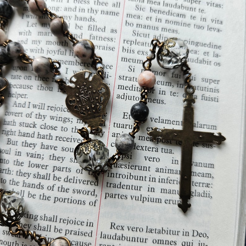 Pu&ograve; includere: Un rosario con una croce d'argento e un ciondolo a forma di cuore. Le perle sono fatte di diversi materiali, tra cui vetro, metallo e pietra. Il rosario &egrave; appoggiato su una pagina di un libro con testo in latino.