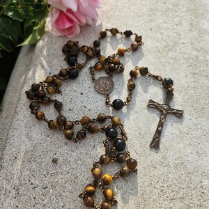 May include: A rosary featuring tiger eye beads and a bronze crucifix. The beads, in shades of brown and black, are linked with bronze wire. A small, circular medal and a pink rose are also present in the image.