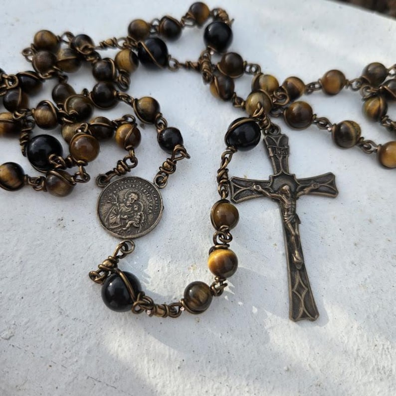 May include: An antique rosary featuring tiger eye and dark brown beads. It includes a metal crucifix and a circular medal with an embossed image. The beads are strung on a metal chain.