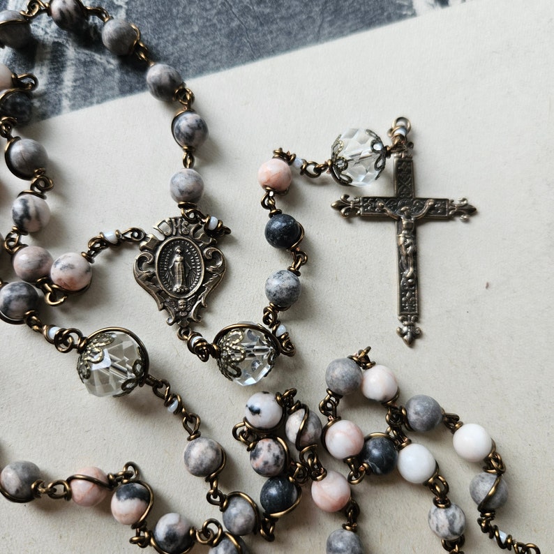 Pu&ograve; includere: Un rosario con perline rosa, grigie e nere, dettagli in cristallo e un crocifisso argentato.