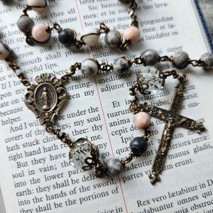 Pu&ograve; includere: Un rosario con perline grigie e rosa, un crocifisso in ottone e una medaglia in ottone con la raffigurazione della Vergine Maria. Il rosario &egrave; disposto su una pagina di un libro con testo in inglese.