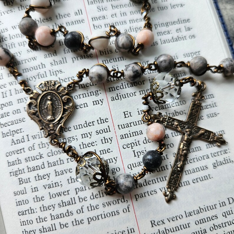 Pu&ograve; includere: Un rosario con perline grigie e rosa, un crocifisso in ottone e una medaglia in ottone con la raffigurazione della Vergine Maria. Il rosario &egrave; disposto su una pagina di un libro con testo in inglese.