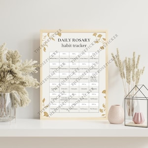 May include: A framed print with the text "DAILY ROSARY habit tracker" in black font. The tracker is a grid with spaces for daily entries. The frame is light-colored, and the print has floral accents in gold.