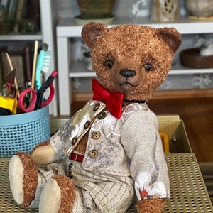 Puede incluir: Un oso de peluche marrón hecho a mano con una chaqueta estampada, pantalones a rayas y una pajarita roja. El oso tiene una cara detallada y está sentado, mostrando su atuendo. El atuendo del oso incluye un cinturón rojo y botones decorativos.