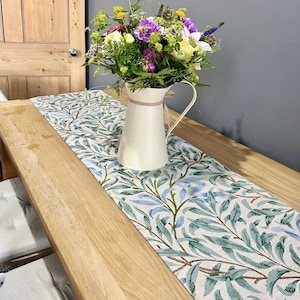 May include: A white table runner with a green and blue floral pattern. The runner is on a wooden table with a white jug filled with flowers.