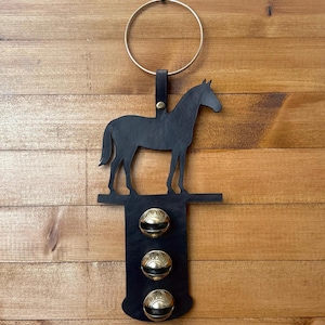 以下が含まれることがあります： 黒い革の馬のシルエット、真鍮のベル3つ、金色のリング。馬はベルの上にある水平なバーの上に立っています。背景は木製の表面です。
