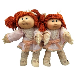 May include: Two dolls with yarn hair in a vibrant orange shade, styled in pigtails with small white bows. They wear matching pale pink dresses featuring lace trim, button details, and a waist ribbon, paired with patterned leggings and cream shoes.