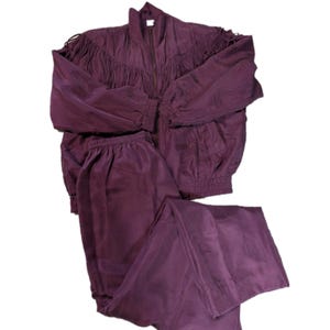 May include: A plum-colored tracksuit set, featuring a zip-up jacket with fringe detailing across the chest and shoulders, and matching pants. The jacket has a high collar and elastic cuffs. The pants have an elastic waistband.