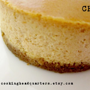 May include: A close-up of a round cheesecake with a golden brown crust. The cheesecake is topped with a smooth, creamy layer of cheesecake filling. The text "CHQ" is visible on the top of the cheesecake.