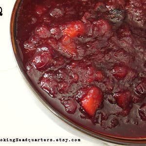 Puede incluir: Un primer plano de un bol de salsa de arándanos. La salsa es de color rojo intenso y tiene una textura lisa y brillante. La salsa está llena de arándanos y algunas pequeñas piezas de cáscara de naranja.