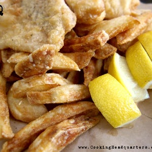 Op de afbeelding: Een close-up van een bord met fish and chips met citroenschijfjes. De vis is gepaneerd en gebakken, en de frietjes zijn goudbruin. De citroenschijfjes zijn heldergeel.