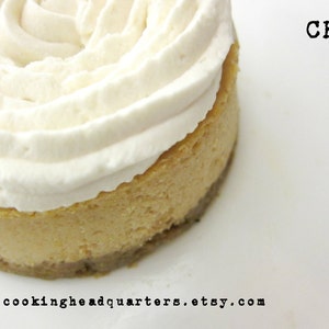May include: A close-up of a small, round cheesecake topped with a swirl of white whipped cream. The cheesecake is on a white plate with the text "CHQ" and "www.cookingheadquarters.etsy.com" printed on the plate.